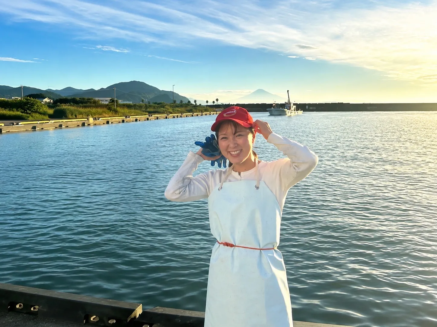 焼津の漁港で、富士山を背に赤い帽子と白い前掛け姿で微笑むやいナビの発信者